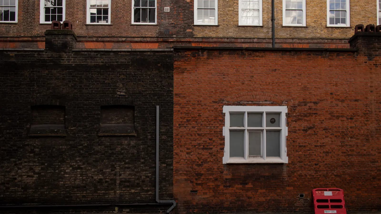 Two brick walls of different ages meeting at a boundary in London, one dark and weathered and one red with a white-framed window