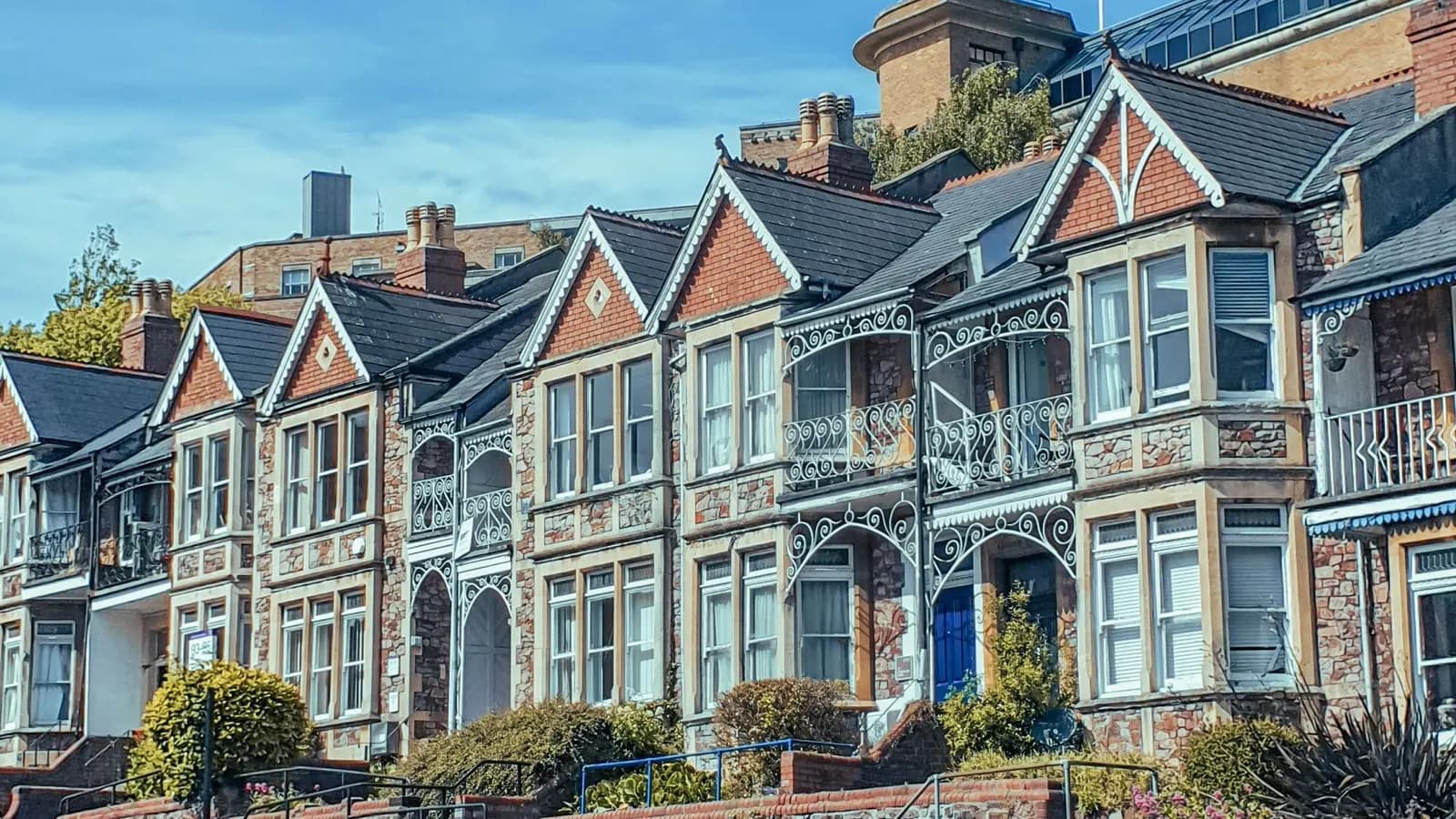 A row of Victorian villas in red brick and pale stone with gables, bay windows and iron balconies, set above a brick garden wall in Bristol
