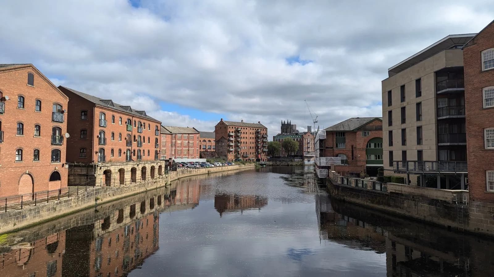 Red brick former warehouses lining a river in central Leeds, with newer buildings on the right and cloud above