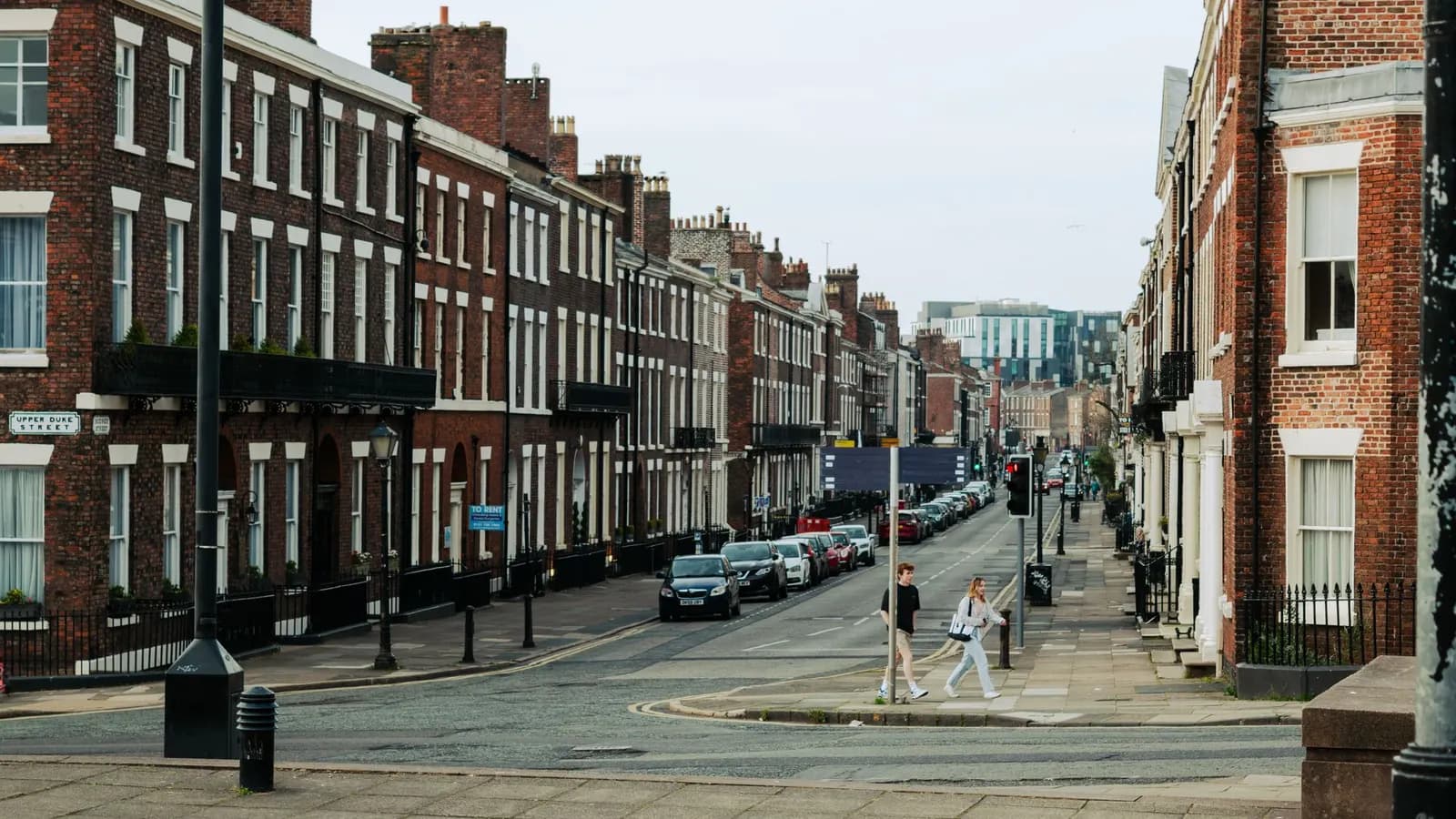 A long Georgian brick street in Liverpool with tall sash windows, iron railings, parked cars and two pedestrians crossing the road