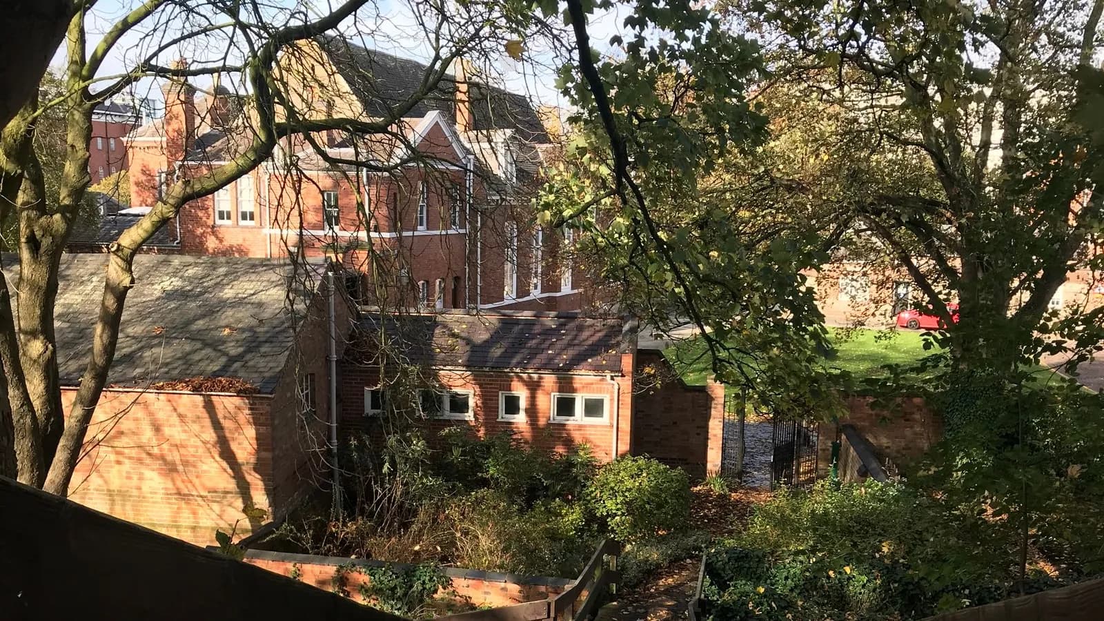 A large Victorian red-brick house with tall chimneys seen over slate-roofed brick outbuildings and autumn trees from a wooden footpath staircase in Leicester