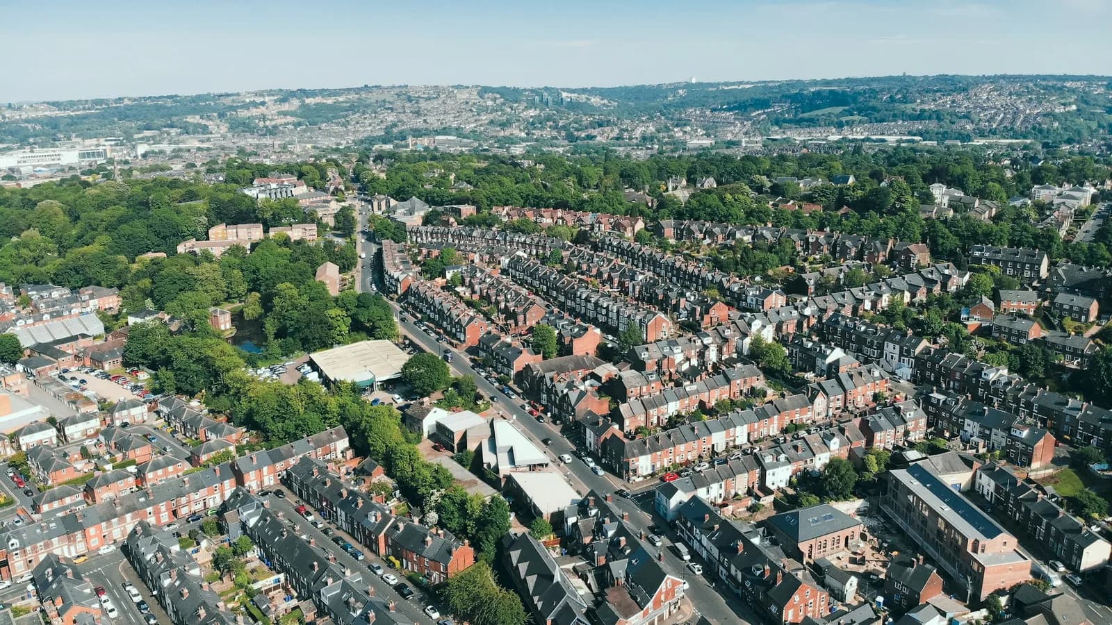 Aerial view over south-west Sheffield showing brick terraced streets, some with roof dormers, a main road and wooded parkland under a clear blue sky