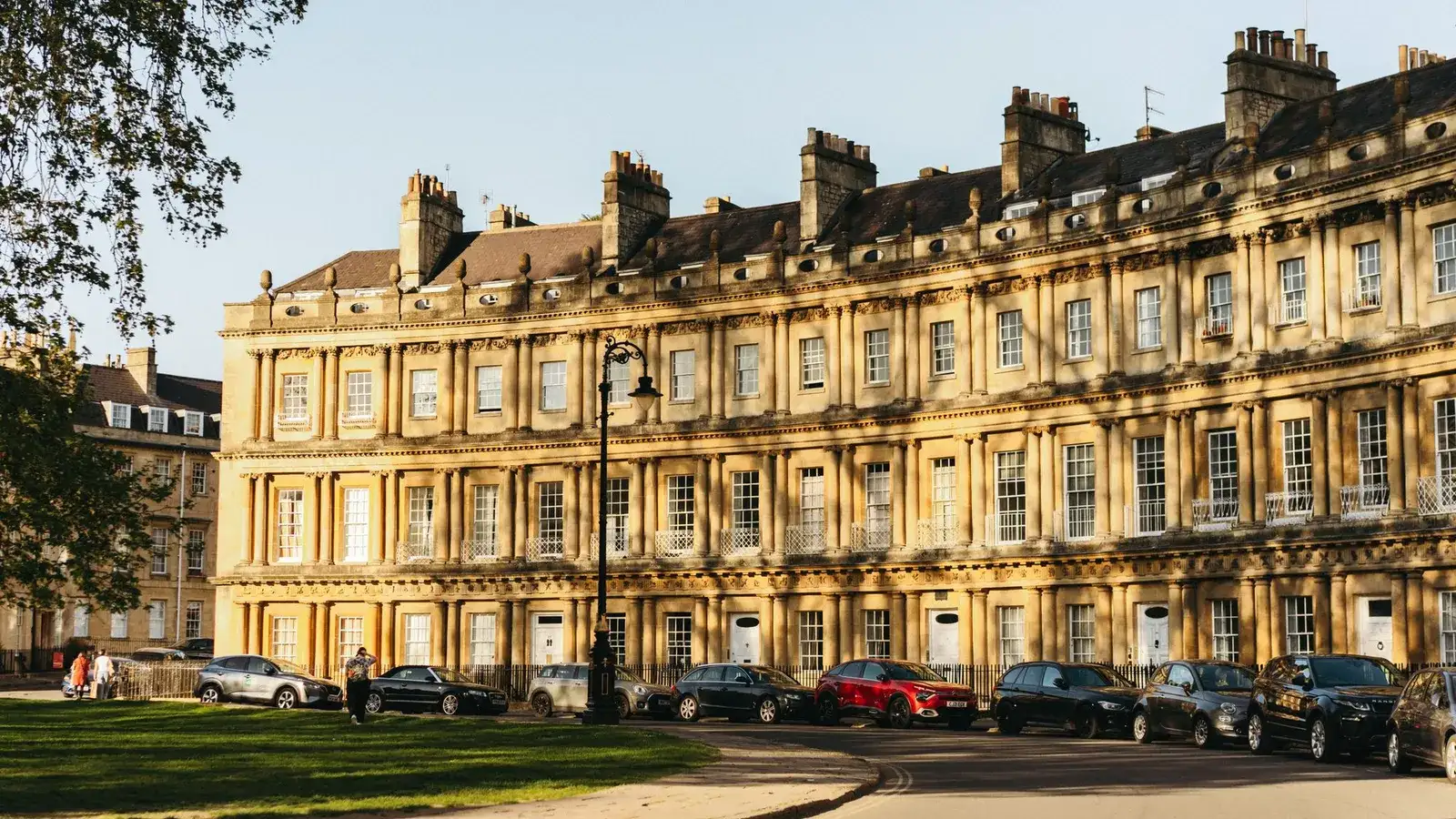 A curved Georgian terrace of honey-coloured Bath stone with three tiers of columns, tall chimney stacks and a slate roof, seen across a green with parked cars
