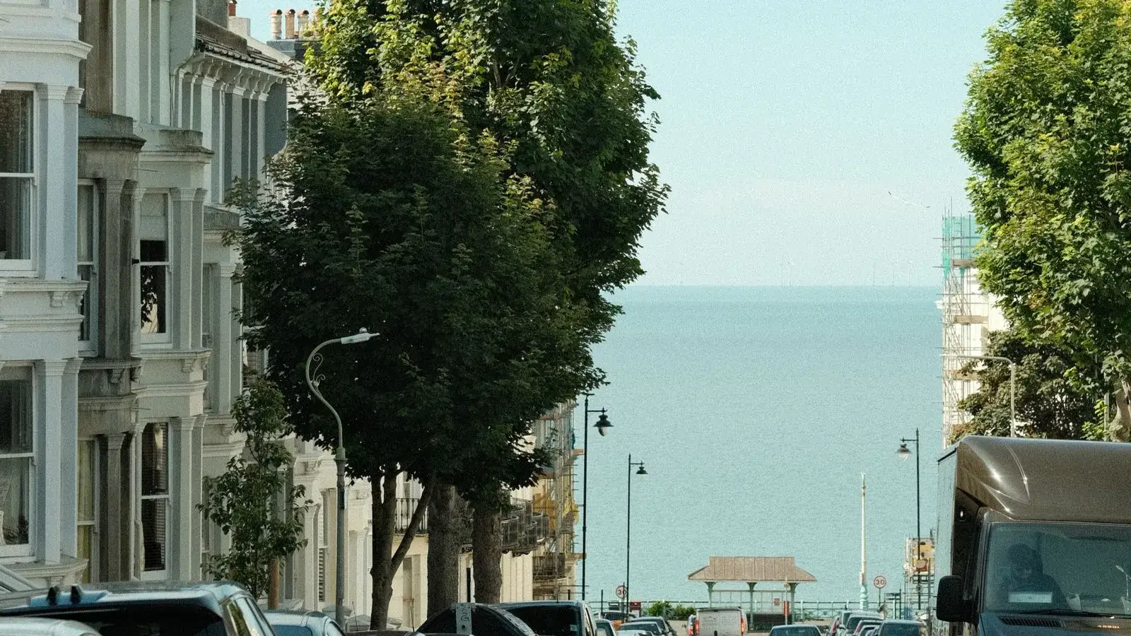 White stucco terraced houses with tall bay windows beside plane trees on a Brighton street that slopes down to the sea, with a seafront shelter at the bottom