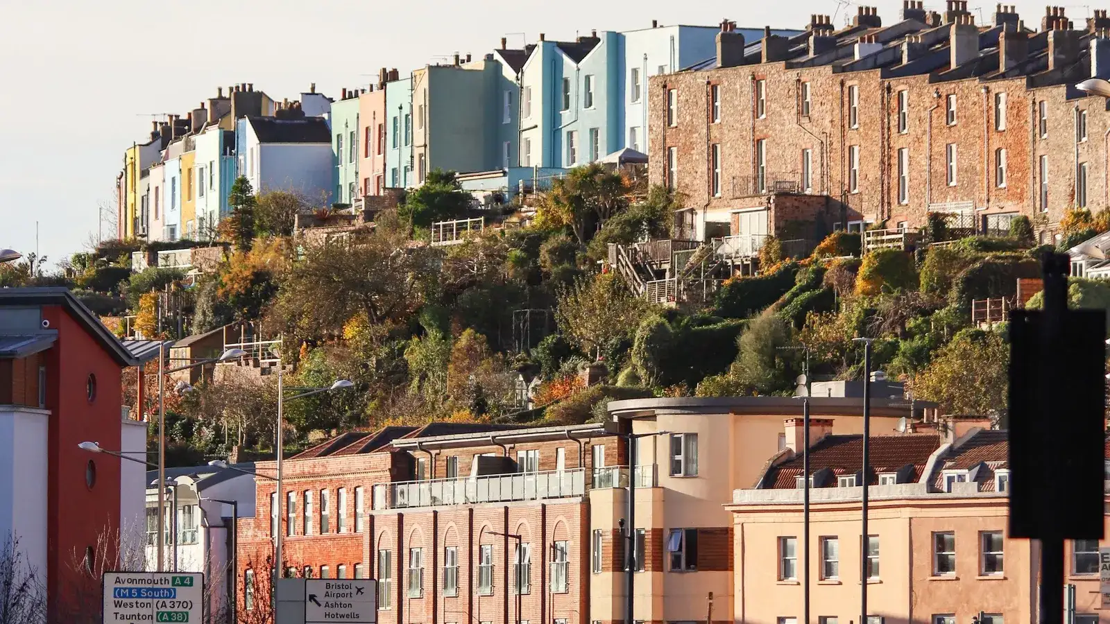 Brightly painted terraced houses and a brick terrace with tall chimneys on a wooded Bristol hillside above a busy main road