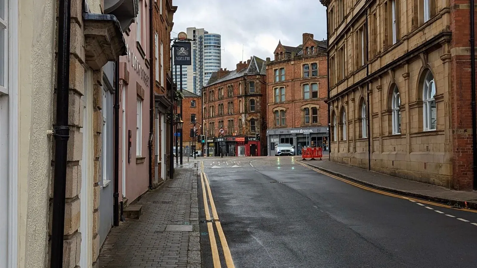 A cobbled Leeds street with sandstone and red brick Victorian buildings, double yellow lines and a modern tower in the distance under a grey sky