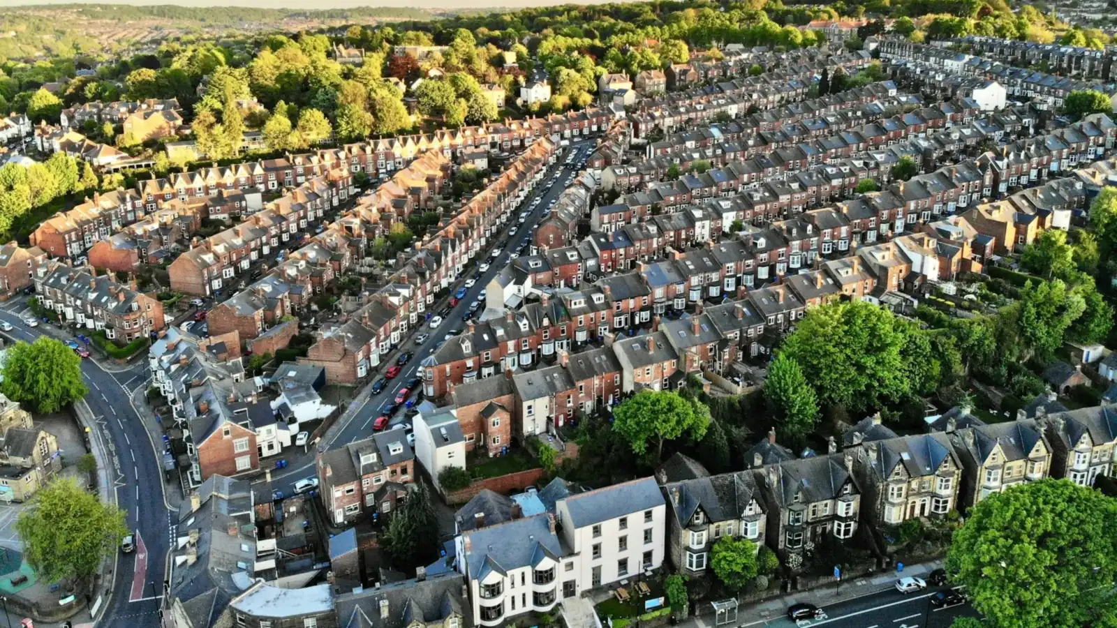 Aerial view of long rows of red brick terraced houses with slate roofs running up a hillside in Sheffield, with a stone-built villa terrace and trees beyond